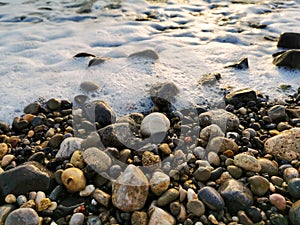 Dark grey, brown, beige, black different size pebbles with selective focus against blurred sea foam background