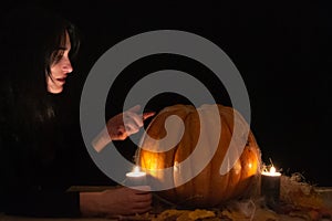 Dark hair pretty woman with big orange pumpkin on black background. The light of the candles illuminates her face. Conceptual