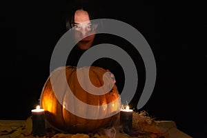 Dark hair pretty woman with big orange pumpkin on black background. The light of the candles illuminates her face. Conceptual