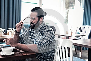 Dark-haired bearded man putting his glasses on before working on laptop