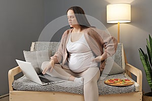 Dark haired Caucasian ill woman wearing casual clothing sitting at home in sofa and working online using laptop touching painful