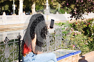A dark-haired, curly-haired adult woman with latin features is sitting on a park bench looking at her mobile phone. Concept of
