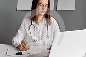 Dark haired female doctor working at office desk wearing glasses and white lab coat looking at laptop screen writing down filling