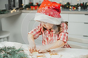 Dark-haired girl 3 years old in red Christmas cap and checkered shirt cuts out gingerbread cookies from rolled dough