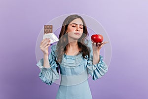 Dark-haired girl in blue dress sadly looks at apple, wanting to eat delicious chocolate bar. Portrait of female model on