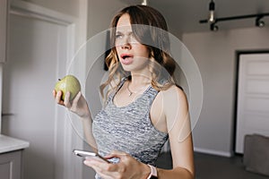 Dark-haired girl with nude makeup posing at home with smartphone. Unhappy young woman with apple an