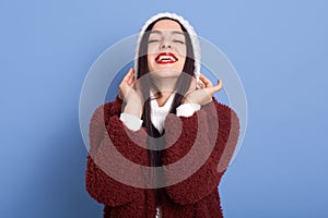 Dark haired girl puts on winter hat over isolated blue background laughing, showing her teeth, wears red lipstick, expressing