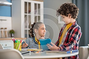 Dark-haired girl sitting at table, writing in her notebook, curly boy standing next to her, showing something on tablet