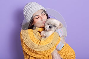 Dark haired girl wearing warm yellow sweater and cap kissing her little dog, blithesome european woman expressing love to her