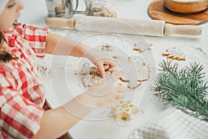 Dark-haired girl 3 years old in red Christmas cap and checkered shirt cuts out gingerbread cookies from rolled dough