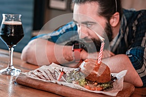 Dark-haired gourmet man looking at glass of beer and meat burger