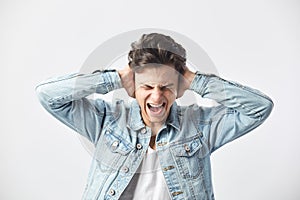Dark-haired guy dressed in a white t-shirt and a denim jacket covers his ears with his hands and shouts on the white