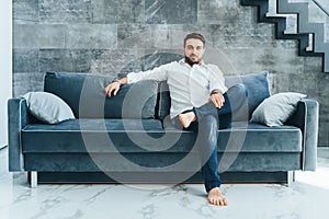 Dark-haired handsome, barefoot confident and smiling bearded businessman looking at camera, sitting on soft sofa indoors