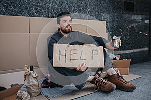 Dark-haired man is sitting on the cardboard and holding a sign that says help. He is holding in left hand a metal cup