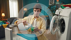 Dark-haired man sitting next to a washing machine, pouring a liquid detergent into a cup to fill the dispenser, planning