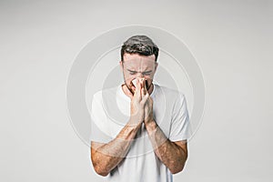 Dark-haired man is standing near the white wall and sneezing. Seems like he caught some cold and soon will be very ill