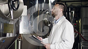 A dark-haired man in a white robe writing down readings of a product in a brewery. A side view slow motion close up