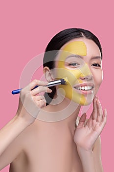 Dark-haired pretty asian girl applying clay facial mask and looking contented
