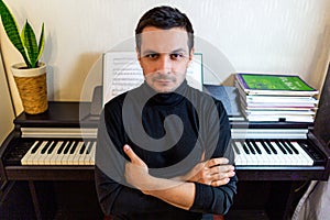 A dark-haired tanned young guy wearing a black turtleneck sitting with arms crossed with his back to the digital piano
