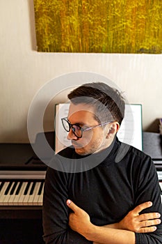 A dark-haired tanned young guy wearing glasses and a black turtleneck sitting with arms crossed with his back to the digital piano