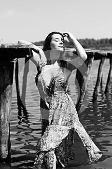 Dark-haired woman posing at the wooden pier at the lake. Woman in summer outfit with her back turned to the wooden footbridge of