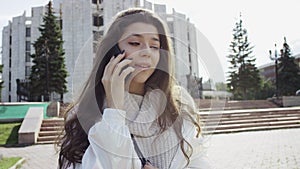 A dark-haired woman in white clothes having a chat with her friend on the smartpohne while standing in the park. A slow