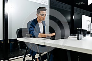 Dark-haired young architect dressed in a blue jacket works on the laptop in the office