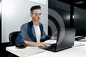 Dark-haired young architect dressed in a blue jacket works on the laptop in the office