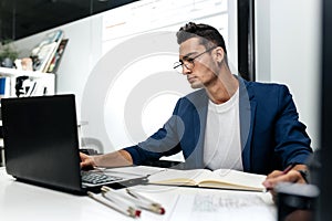 Dark-haired young architect dressed in a blue jacket works on the laptop in the office