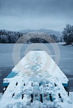 Tmavá krajina zvislá fotografia mola pokrytého snehom so zamrznutým jazerom na pozadí - zimný čas. Zamrznuté a studené jazero