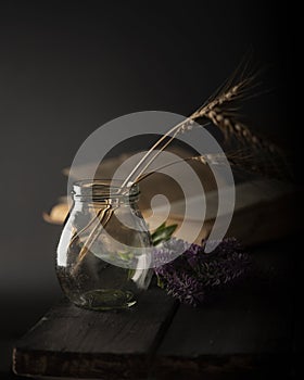 Dark moody still life with book, flower and glass bottle with ears of wheat on wooden table.