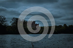 Dark and moody view of castle Svaneholms slott embedded in forest beyond lake on a stormy spring evening