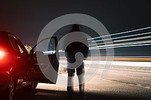 A dark mysterious blurred figure standing on a road, next to car. Next to traffic trails on a country road at night