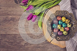 Dark photo of Easter quail eggs in an ancient metal bowl. Beautiful spring flowers - purple tulips on a wooden background. Floral