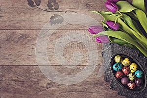 Dark photo of Easter quail eggs in an ancient metal bowl. Beautiful spring flowers - purple tulips on a wooden background. Floral