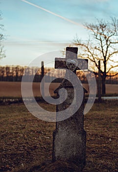 Dark photo of old and abandoned cross grave stone on European cemetery with tree and forest on background on sunset. Creepy and