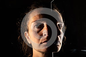 Dark portrait of a serious woman with only half her face illuminated on a black background that is unfolded in the reflection of a