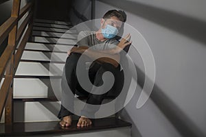Dark portrait of young scared and worried man in protective mask sitting on stairs at home staircase during lockdown and photo