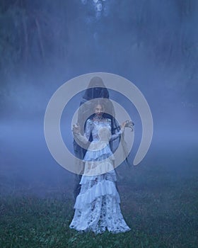 Dark Queen with neat hairdo in a white vintage dress and a silver necklace, standing in a forest full of thick purple