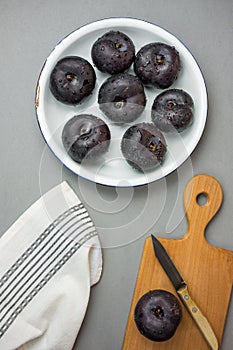 Dark red big plums on white vintage enamel plate. Wooden cutting board knife white cotton towel on gray stone. Minimalist