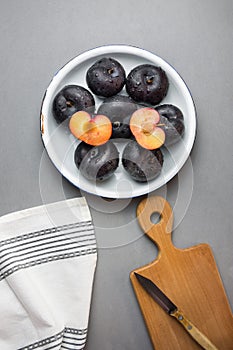 Dark red big plums whole and halved on white vintage enamel plate. Wooden cutting board knife white cotton towel on gray stone