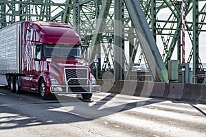 Dark red big rig bonnet semi truck transporting cargo in refrigerator semi trailer driving on the arched truss bridge