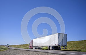 Dark red big rig semi truck transporting commercial cargo in dry van semi trailer driving on the narrow road with hill on the side