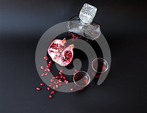 Dark red liqueur in two glasses and a glass bottle on a black matte background next to the seeds and pomegranate fruit