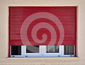 Dark red rolling shutters and white frame window on beige wall,