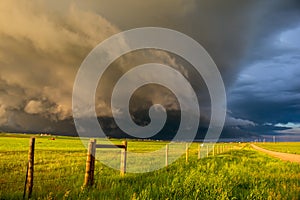 A dark shelf cloud and storm approach as the sun shines brightly looking down a fence.