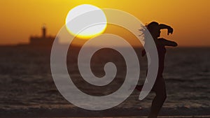 Dark silhouette of dancing young woman on the seashore during bright orange sunset