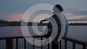Dark silhouette of dreamy woman sitting on wooden rail of footbridge on lake at sunrise. Female person enjoying nature