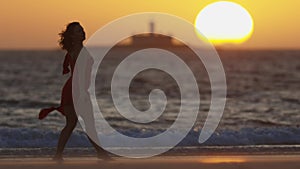 Dark silhouette of graceful young woman dancing on the seashore during bright orange sunset