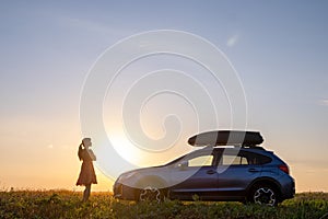 Dark silhouette of lonely woman relaxing near her car on grassy meadow enjoying view of colorful sunrise. Young female